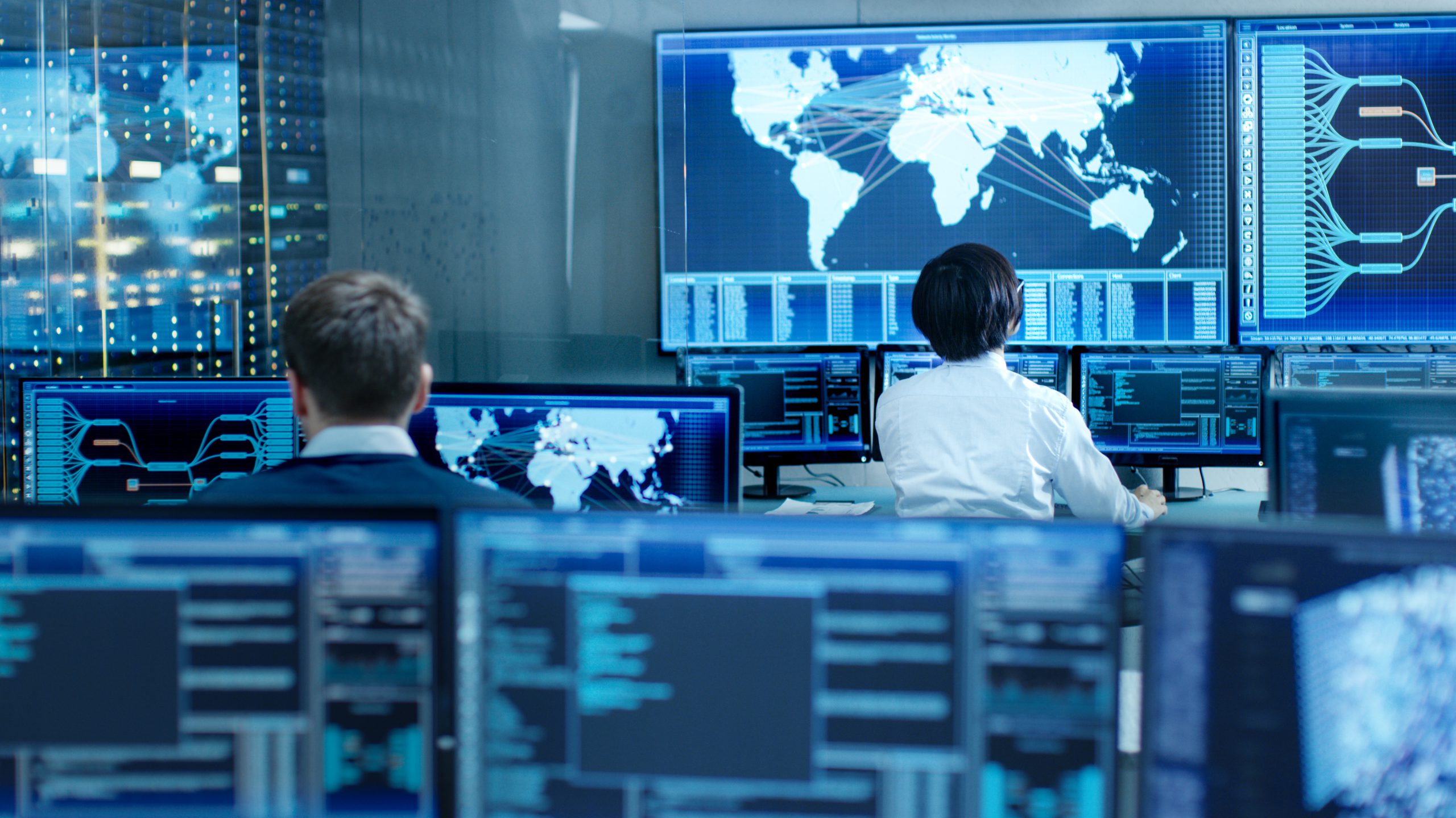 In the System Control Room Operator and Administrator Sitting at Their Workstations with Multiple Displays Showing Graphics and Logistics Information.